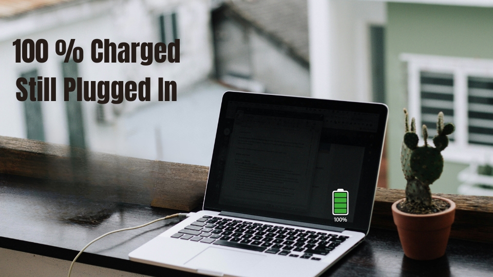 A laptop sitting on a wooden ledge outdoors, plugged into a charger with a full 100% green battery icon overlay and text reading "100% Charged Still Plugged In".